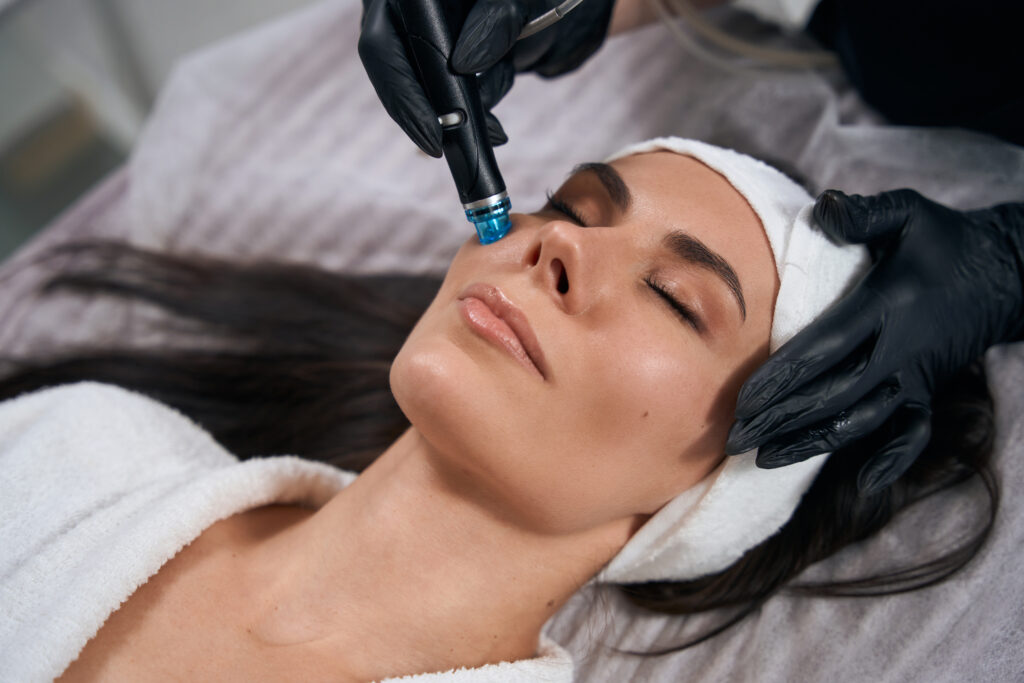 Cosmetologist in protective gloves sitting near adult woman and doing HydraFacial procedure in office