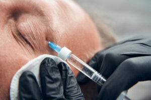 Close up of doctor in sterile gloves using syringe and cotton pad while making injection in man face